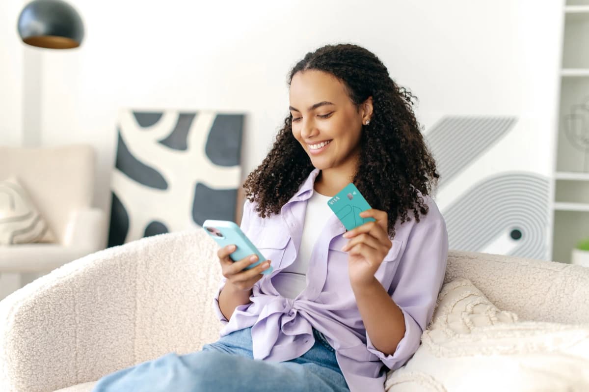 Na imagem você vê uma jovem negra com um belo cabelo cacheado e um belo sorriso enquanto segura o METTAcard em uma mão e acessa o aplicativo com a outra utilizando o celular